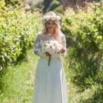 Uma mulher entre fiadas de videiras verdes, com o crop top Madeira branco, de mangas compridas transparentes e renda floral, sobre a Saia Anadia fluida. Olha para baixo, para um ramo de rosas cor de creme, com uma coroa de flores a condizer, debaixo de uma palmeira.