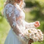Uma mulher com o crop top Madeira branco, de mangas compridas transparentes e renda floral detalhada, sobre a Saia Anadia fluida. Olha para baixo, a segurar um ramo de rosas cor de creme, com uma coroa de flores a condizer num jardim ao sol.