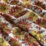 Floral jacquard fabric in overlapping folds, featuring large rust red, coral pink and golden yellow flowers with dark grey leaves on an ivory background and shimmering gold-toned details.