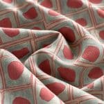 Geometric fabric arranged in soft overlapping folds, featuring irregular coral pink shapes inside a grid of fine coral double-lined squares on a silver grey background.