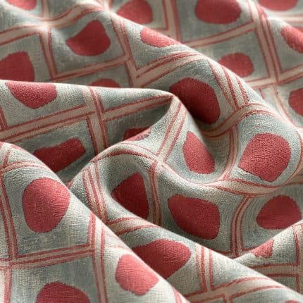 Geometric fabric arranged in soft overlapping folds, featuring irregular coral pink shapes inside a grid of fine coral double-lined squares on a silver grey background.
