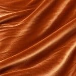 Copper orange crystal velvet fabric arranged in broad diagonal folds, revealing its smooth pile and reflective sheen.