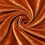 Copper orange crystal velvet fabric arranged in a spiral swirl, showing the plush texture, rich colour and soft flowing drape.