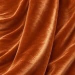 Copper orange crystal velvet fabric draped in soft vertical folds, highlighting its smooth surface, fluid drape and luminous sheen.