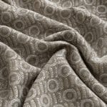 Close-up of sage green and silver grey jacquard fabric draped in loose folds, highlighting the textured concentric circles, angular woven details and alternating light and shadow.