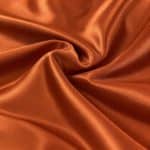 Copper orange Moroccan sablé fabric gathered into soft curved folds around a loose central swirl, showing its subtly textured surface and bright, satin-like highlights.