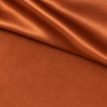 Copper orange Moroccan sablé fabric shown flat on its softly textured matte side beside a folded, smooth satin side with bright reflective highlights, revealing the contrast between the two finishes.