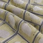 Soft yellow, grey and beige geometric jacquard arranged in overlapping folds, showing raised pale oval motifs inside grey-outlined squares.