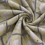Geometric jacquard fabric gathered into a tight central spiral, with soft yellow squares, grey grid lines and irregular pale beige ovals curving through the folds.