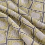 Soft yellow and grey geometric jacquard draped in broad diagonal folds, revealing textured beige oval motifs, grey borders and dark flecked details.