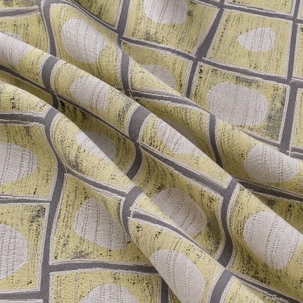 Soft yellow and grey geometric jacquard draped in broad diagonal folds, revealing textured beige oval motifs, grey borders and dark flecked details.