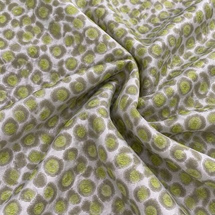 Silver-grey silk jacquard fabric gathered in soft folds, featuring repeating irregular circular motifs with textured lime green centres and grey-green outlines.