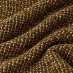 Textured wool fabric arranged in overlapping folds, with tightly clustered olive green, cream and brown yarn loops creating a raised, speckled surface on a dark brown ground.